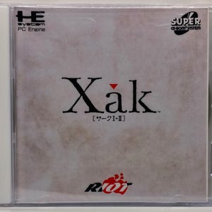 May include: A PC Engine game case for "Xak". The cover features the title in a stylized font with a red accent mark. The case has the "SUPER CD-ROM SYSTEM" logo and the "RIOT" logo. The game title is also written in Japanese.