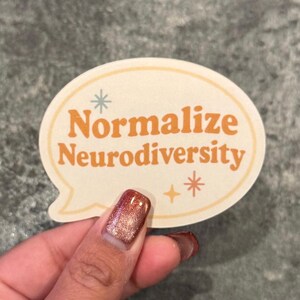 May include: A sticker with the text "Normalize Neurodiversity" in orange, within a cream-colored speech bubble shape. The sticker has small star and asterisk designs. The image shows a hand holding the sticker.