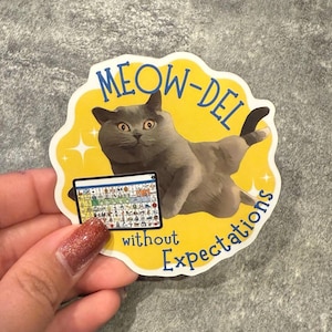 May include: A sticker featuring a gray cat in a relaxed pose, with the words "MEOW-DEL" and "without Expectations" in yellow. The cat is holding a small tablet with colorful icons. The sticker has a yellow border with star accents.