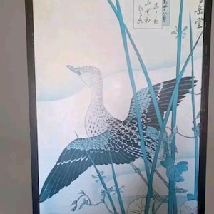 Könnte beinhalten: Gerahmter japanischer Holzschnitt mit einem Vogel mit ausgebreiteten Flügeln in Blau-, Schwarz- und Weißtönen. Das Kunstwerk zeigt stilisierte Wasserpflanzen und japanischen Text vor einem hellblauen Hintergrund.