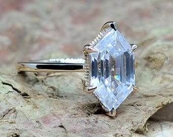 May include: A gold ring with a large, clear, hexagonal-cut gemstone. The gemstone is the focal point, reflecting light. The ring's band is thin and gold, with delicate detailing near the gemstone setting. The ring is set on a textured surface.