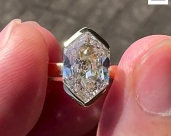 May include: A gold ring featuring a large, hexagonal-cut diamond. The diamond is clear and multifaceted, reflecting light. The ring is held between two fingers, showcasing the intricate design and sparkle of the gemstone.