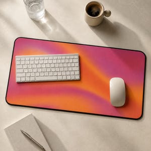 May include: A rectangular desk mat featuring a vibrant gradient of pink, magenta, and orange hues with a smooth surface. A white wireless keyboard and a white computer mouse are positioned on top of the mat, which has a thin black border.