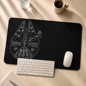 May include: A rectangular black desk mat featuring a white line art illustration of the Millennium Falcon spaceship against a starry background. The mat is positioned under a white computer keyboard and a white computer mouse.
