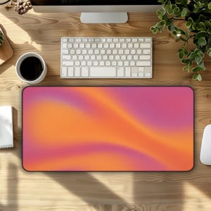 May include: A rectangular mouse pad with a vibrant gradient design in shades of orange and pink. The pad has a black border and is placed on a wooden desk, alongside a white keyboard, a coffee cup, and a computer mouse.