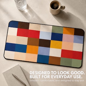 May include: A rectangular desk mat featuring a grid pattern of solid color blocks in shades of blue, red, yellow, green, brown, beige, and white. The mat has a black border and includes the text: DESIGNED TO LOOK GOOD. BUILT FOR EVERYDAY USE.