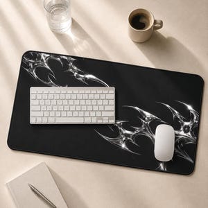 May include: A rectangular black desk mat featuring a metallic silver abstract tribal pattern. The mat is positioned under a white wireless keyboard and a white computer mouse. The design consists of sharp, fluid, chrome-like shapes that contrast against the solid black background of the mat surface.