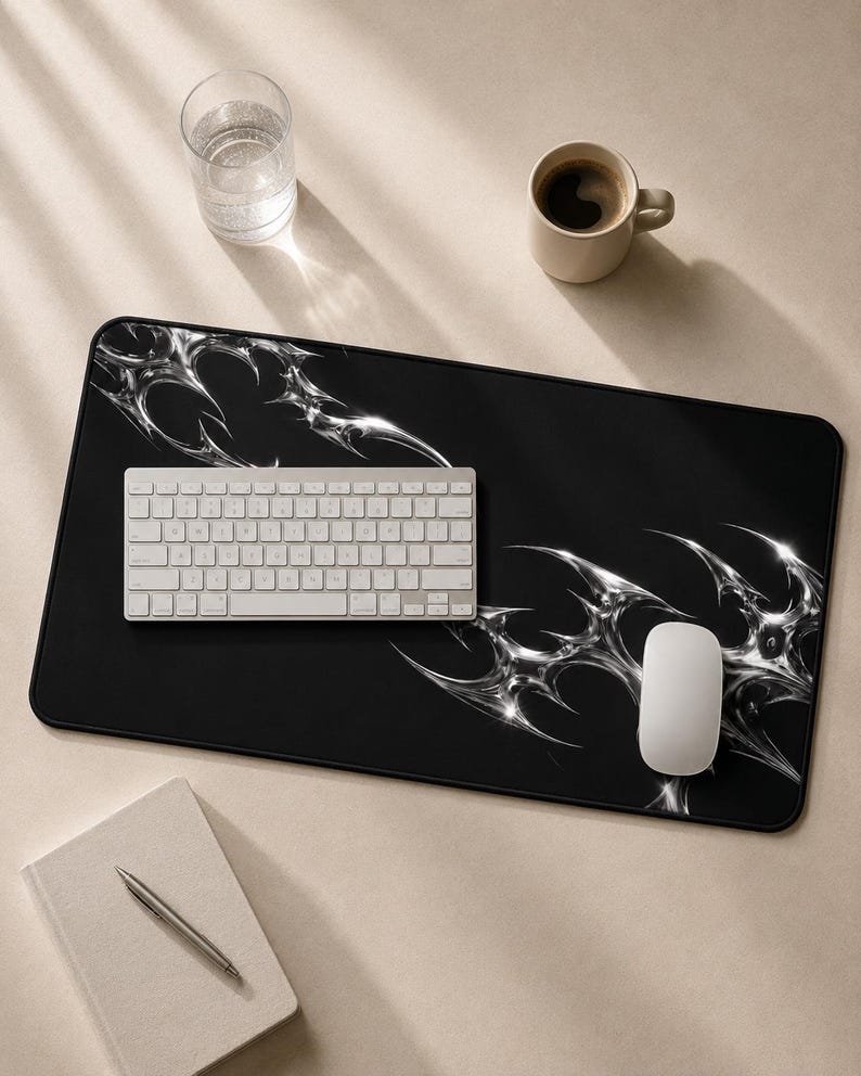 May include: A rectangular black desk mat featuring a metallic silver liquid-like abstract pattern in the corners. The mat is positioned under a white wireless keyboard and a white computer mouse, providing a smooth surface for office equipment.