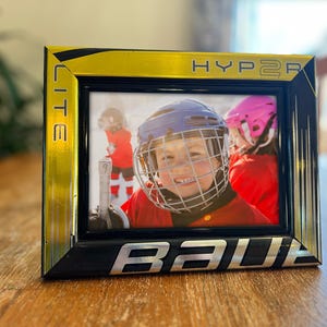May include: A black and gold picture frame with the words "HYPERLITE" and "BAUER" on it. The frame has a photo of a young hockey player wearing a helmet and a red jersey.
