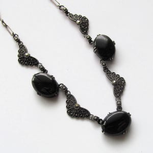 May include: A vintage-style necklace featuring oval black stones and ornate, dark metal filigree accents. The necklace has a delicate chain and is designed with a symmetrical pattern. The black stones are smooth and polished, contrasting with the detailed metalwork.