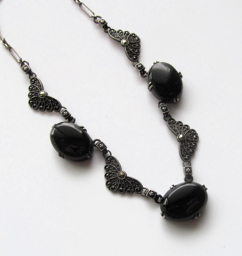 May include: A vintage-style necklace featuring oval black stones and ornate, dark metal filigree accents. The necklace has a delicate chain and is designed with a symmetrical pattern. The black stones are smooth and polished, contrasting with the detailed metalwork.