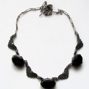 May include: A vintage-style necklace featuring a dark silver chain and decorative elements. The necklace has three large, oval, black gemstones and ornate filigree accents. The chain is connected with small, rectangular links.