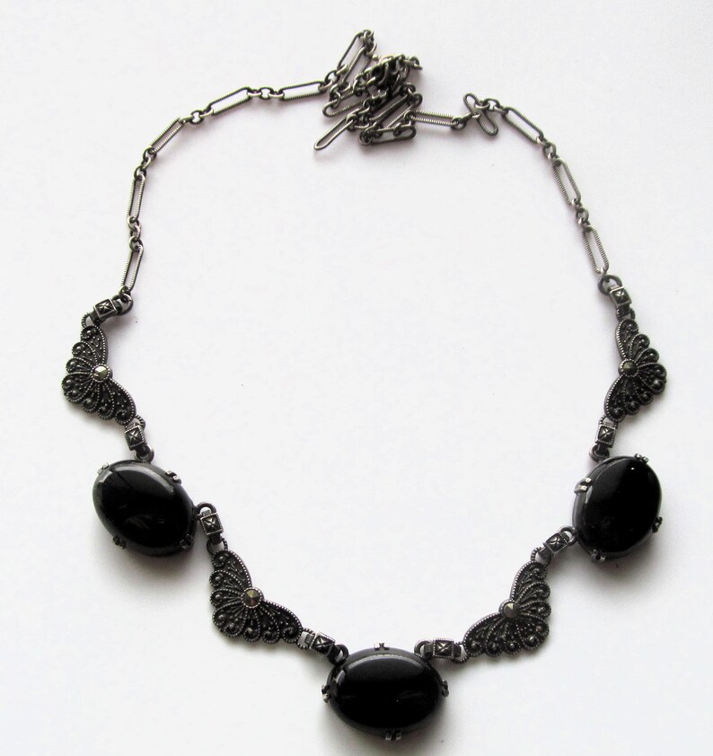 May include: A vintage-style necklace featuring a dark silver chain and decorative elements. The necklace has three large, oval, black gemstones and ornate filigree accents. The chain is connected with small, rectangular links.