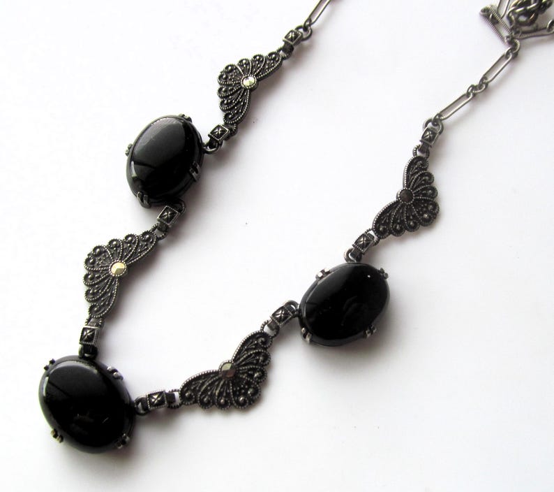 May include: A vintage-style necklace featuring oval black gemstones and ornate silver-tone metalwork. The necklace has a delicate chain and decorative filigree elements, creating an elegant and classic look. The gemstones are set in a bezel setting.