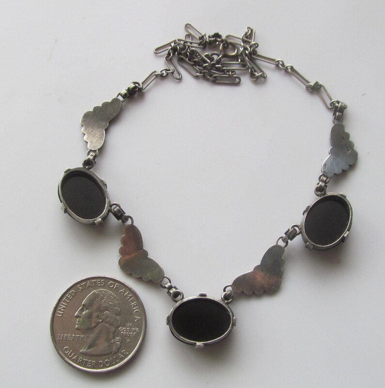 May include: Silver-toned necklace featuring oval black stones and butterfly-shaped accents. The necklace is displayed with a U.S. quarter for size comparison. The chain is delicate and the design is vintage.