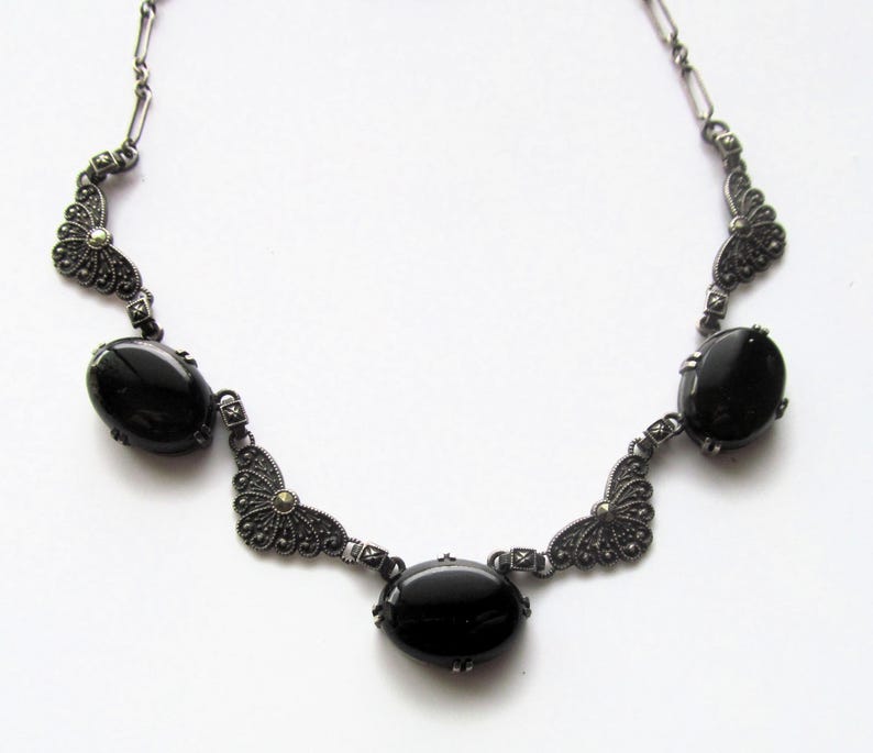 May include: A vintage-style necklace featuring oval black gemstones set in an ornate, dark metal filigree design. The necklace has a symmetrical arrangement with three prominent central stones and decorative accents.