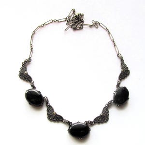 May include: A silver-toned necklace featuring five oval, black gemstones. The necklace has an ornate design with filigree detailing and a delicate chain. The gemstones are evenly spaced along the necklace's length, creating a symmetrical and elegant look.