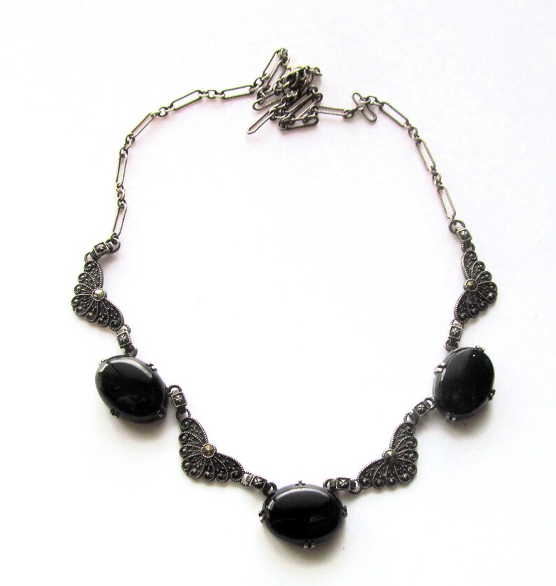 May include: A silver-toned necklace featuring five oval, black gemstones. The necklace has an ornate design with filigree detailing and a delicate chain. The gemstones are evenly spaced along the necklace's length, creating a symmetrical and elegant look.