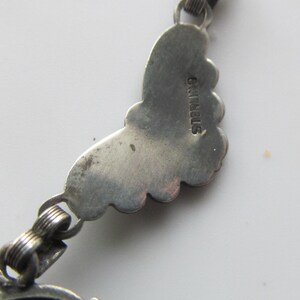 May include: Close-up of a silver-toned jewelry piece. The piece features a decorative, scalloped edge design. The word "STERLING" is stamped on the back of the piece. The jewelry is connected with small, looped links.