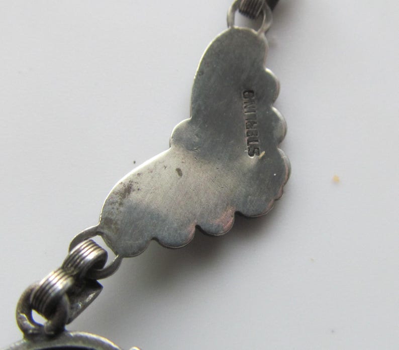 May include: Close-up of a silver-toned jewelry piece. The piece features a decorative, scalloped edge design. The word "STERLING" is stamped on the back of the piece. The jewelry is connected with small, looped links.