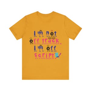 Peut inclure: Un t-shirt à manches courtes jaune moutarde avec le texte I'm not off track. I'm off script. Les lettres sont stylisées avec de petites jambes et bras de bonhomme allumette, incluant des icônes comme des couronnes et des boutons d'alimentation.