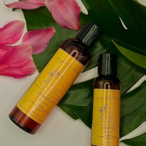 May include: Two brown glass bottles of Island Girl Body Oil with black caps, one 4 fl oz and one 2 fl oz. The bottles have yellow labels with a palm tree and sunset design. Pink flower petals and a green leaf are in the background.