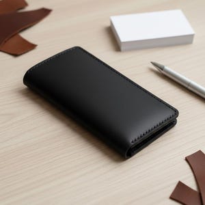 May include: A black leather wallet with stitched edges sits on a light wood surface. A stack of white cards and a silver pen are nearby. Brown leather offcuts are also visible, suggesting a handcrafted item.