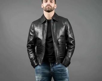 May include: A black leather jacket with a crocodile skin texture. The jacket has a zipper closure, two front pockets, and a collared neckline. The person in the image is wearing blue jeans and a black shirt.