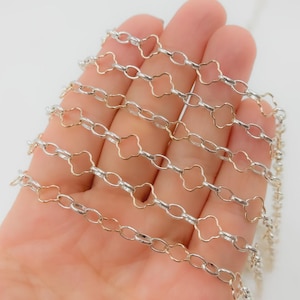 May include: A close-up of a silver and gold chain. The chain features a repeating pattern of silver oval links connected by gold clover-shaped links. The chain is displayed against a light background.