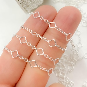 May include: Close-up of a silver chain with a repeating geometric diamond-shaped design. The chain is displayed against a light background, highlighting its intricate details and reflective surface. The chain is likely used for jewelry making.