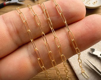 May include: A gold-toned paperclip chain featuring elongated rectangular links. The delicate metal chain is held in place, showcasing its uniform structure and polished finish, suitable for jewelry making or as a standalone necklace component.