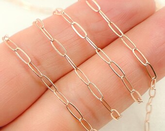 May include: A delicate gold chain with elongated oval links. The chain is a warm, rose gold color and is displayed against a blurred background. The links are evenly spaced, creating a simple yet elegant design. This chain is suitable for necklaces or bracelets.