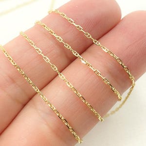 May include: Close-up of a delicate gold chain with a repeating anchor link design. The chain is a bright, shiny gold color and appears to be lightweight. The chain is held by a person's fingers.