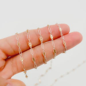 May include: Close-up of a delicate gold and silver chain necklace. The chain features alternating oval and diamond-shaped links, with a two-tone design. The necklace is displayed against a light background.