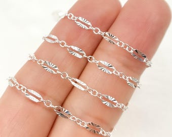 May include: Close-up of a delicate silver chain with oval-shaped links. Each link has a textured, patterned design. The chain is displayed against a light background, highlighting its intricate details. Ideal for jewelry making.