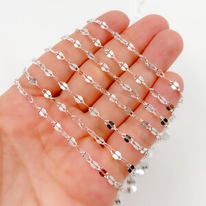 May include: A delicate silver chain featuring a repeating pattern of small, flat, oval links. The chain is held in a hand, highlighting its reflective surface and intricate design. The chain is suitable for jewelry making.