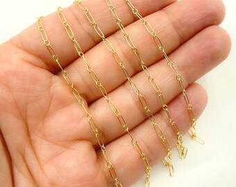 May include: A delicate gold-colored chain necklace with an elongated oval link design. The necklace is held in a hand, showcasing its fine details and subtle shine. The chain is a simple yet elegant piece of jewelry.