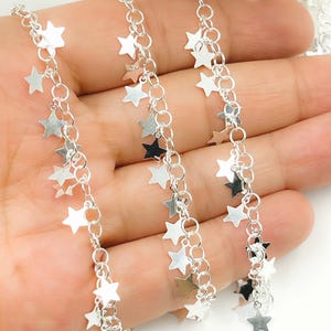 May include: Close-up of silver-colored chain with dangling star charms. The chain is composed of small, interconnected rings, and the stars are of varying sizes. The background is a hand, suggesting the chain is being displayed or worn.