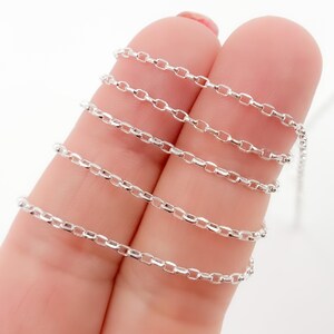 May include: A close-up of a delicate silver chain with small, oval links. The chain is draped across two fingers, showcasing its fine details and reflective surface. The chain is a bright, shiny silver color.