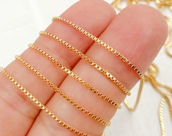 May include: Close-up of several strands of gold-coloured box chain necklaces. The chains are displayed against a light background, showcasing their delicate, square-link design. The chains are tightly woven, reflecting light and highlighting their metallic sheen.