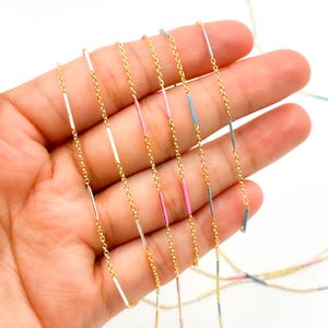 May include: Delicate gold-tone necklaces with colorful bar pendants. The necklaces feature thin gold chains with small, vertical bars in white, pink, blue, and gray. The jewelry is displayed against a light background.
