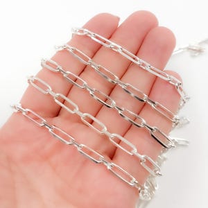 May include: A silver chain featuring elongated oval links. The chain is held in a hand, with the links evenly spaced. The chain is made of a shiny, reflective metal, and the links are smooth and polished. This is a piece of jewellery.