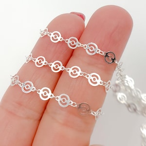 Puede incluir: Primer plano de una delicada cadena de plata con un diseño de círculos abiertos repetidos. La cadena se sostiene entre los dedos, mostrando el intrincado patrón y la superficie reflectante. La cadena es una joya.