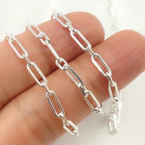 Puede incluir: Un collar de cadena plateada con eslabones ovalados alargados. La cadena se sostiene en una mano, mostrando su superficie brillante y reflectante. Los eslabones están conectados en un estilo clásico de clip, creando un diseño simple pero elegante.