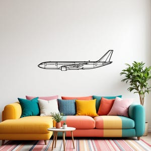 Può includere: Un disegno a tratto nero di un aereo è montato su una parete bianca sopra un divano colorato. Il divano ha una chaise longue gialla e una seduta multicolore con vari cuscini. Un tavolino rotondo e una pianta in vaso sono di fronte al divano.