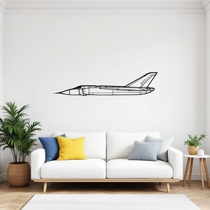 May include: Black line drawing of a jet airplane mounted on a white wall. The artwork is a minimalist depiction of an aircraft, featuring a pointed nose, a streamlined body, and a distinctive tail fin. The jet is displayed above a white sofa with colorful pillows.