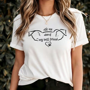May include: White t-shirt with rolled sleeves featuring a black graphic design. The design includes dog ears, a heart, a paw print, and the text "ask me about my best friend."