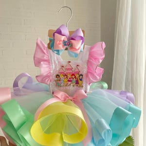 Puede incluir: Un vestido tutú de colores pastel con una diadema a juego. El vestido presenta un corpiño con un gráfico de princesa, mangas con volantes y capas de tul y cintas en tonos rosa, amarillo, verde y azul. Un gran lazo adorna la cintura.
