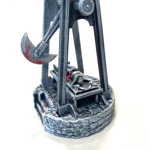 Puede incluir: Un modelo en miniatura de un instrumento de tortura medieval, un potro con una figura tendida sobre él, pintado en un esquema de colores gris y blanco con acentos rojos. La figura está atada al potro y tiene una expresión de angustia en su rostro. El potro está hecho de metal y tiene un hacha grande unida a él.