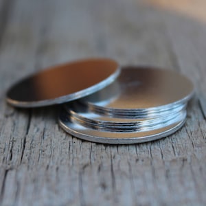 Puede incluir: Una pila de discos de metal de color plateado, probablemente para manualidades o la fabricación de joyas, sobre una superficie de madera.